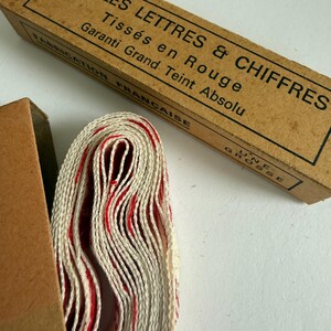 May include: A roll of off-white fabric tape with red woven lettering and numbers, partially inside a rectangular cardboard box. The box features black text reading INITIALES LETTRES & CHIFFRES, Tiss&eacute;s en Rouge, Garanti Grand Teint Absolu.