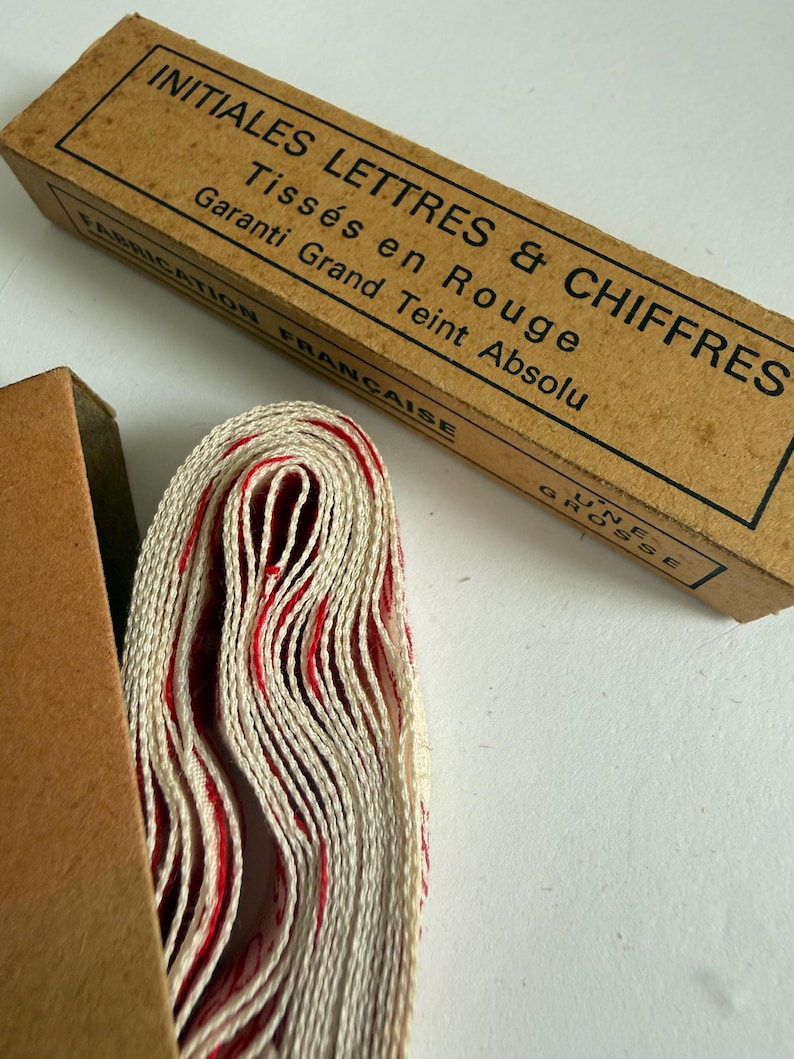 May include: A roll of off-white fabric tape with red woven lettering and numbers, partially inside a rectangular cardboard box. The box features black text reading INITIALES LETTRES & CHIFFRES, Tiss&eacute;s en Rouge, Garanti Grand Teint Absolu.