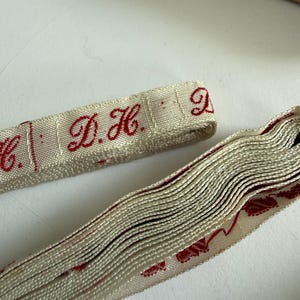 May include: A roll of off-white woven fabric tape featuring repeating red embroidered lettering that reads D.H. The tape has a textured surface and is displayed alongside a flat segment of the same material, showcasing the red monogram detail.