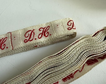May include: A roll of off-white woven fabric tape featuring repeating red embroidered lettering that reads D.H. The tape has a textured surface and is displayed alongside a flat segment of the same material, showcasing the red monogram detail.