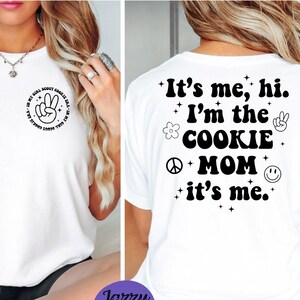 May include: White t-shirt with a peace sign and the text "In my girl scout cookie era, in my girl scout cookie era" on the front. The back of the shirt has the text "It's me, hi. I'm the cookie mom. It's me." with peace signs, flowers, and smiley faces.
