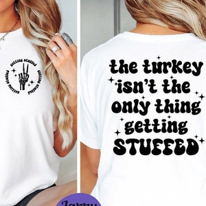 Puede incluir: Camiseta blanca con un signo de paz de mano de esqueleto y el texto "getting stuffed" en la parte delantera. La parte posterior de la camiseta tiene el texto "the turkey isn't the only thing getting stuffed".