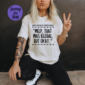 Puede incluir: Camiseta blanca con el texto negro "ME WHILE DRIVING: WELP, THAT WAS ILLEGAL, BUT OKAY." La camiseta tiene cuello redondo y mangas cortas. La marca "Jazzy Joe Customs" está en un círculo morado en la esquina superior izquierda.