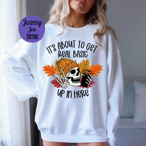 May include: White sweatshirt with a skeleton wearing a beanie and holding a coffee cup. The text on the sweatshirt reads "It's about to get real basic up in here".