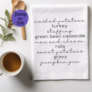 May include: White kitchen towel with black text listing traditional Thanksgiving foods: mashed potatoes, turkey, stuffing, green bean casserole, mac and cheese, rolls, sweet potatoes, gravy, and pumpkin pie.