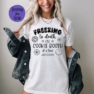 May include: White t-shirt with black text that reads "Freezing to death *ONE* COOKIE BOOTH *at a time* #GIRLSCOUTLIFE". The text is surrounded by snowflakes and a graphic of a Girl Scout cookie.