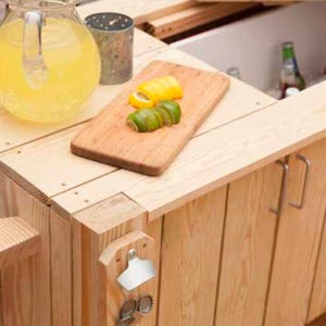 Könnte beinhalten: Ein Holz-Barwagen mit einem Kühlfach, einem Schneidebrett mit Zitronen- und Limettenscheiben, einem Krug Limonade und einem Flaschenöffner. Der Wagen hat ein Handtuch an der Seite und zwei Metallgriffe.