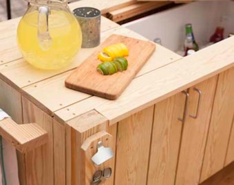 May include: A wooden bar cart with a cooler compartment, a cutting board with lemon and lime slices, a pitcher of lemonade, and a bottle opener. The cart has a towel hanging on the side and two metal handles.