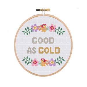 Könnte beinhalten: Ein weißer Stickrahmen mit einem Kreuzstich-Design mit einem floralen Rand und dem Text "GOOD AS GOLD" in goldener Farbe.