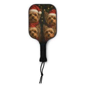 May include: A pickleball paddle featuring a festive design with four Yorkshire Terriers wearing Santa hats and red scarves. The paddle has a black handle with a black cord. The background includes a Christmas tree and twinkling lights.