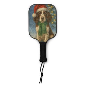 May include: A pickleball paddle featuring a painting of a dog wearing a Santa hat and green scarf. The dog is brown and white with a snowy background and a Christmas tree. The paddle has a black handle with a black cord.