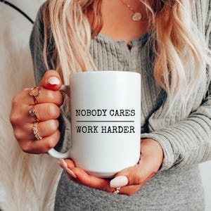 May include: White ceramic mug with the text "NOBODY CARES WORK HARDER" in black. The mug is held by a person wearing rings and a gray long-sleeve shirt. The mug is a standard size for coffee or tea.