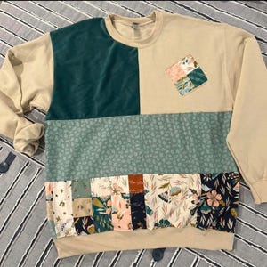 Könnte beinhalten: Ein langärmeliges Sweatshirt mit Rundhalsausschnitt und einem Color-Block-Design in Beige und Dunkel-Türkis. Der untere Bereich zeigt ein Patchwork-Muster aus Blumen- und Pflanzenmotiven in den Farben Rosa, Grün, Marineblau und Creme.