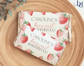 May include: Two white chocolate bar wrappers with a strawberry pattern. The text on the wrappers reads "Caroline's berry first birthday!"