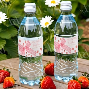 May include: Two clear plastic water bottles with white caps and labels. The labels feature a pink gingham bow, strawberries, and text that reads "Victoria & Robert's Baby Shower". Fresh strawberries are scattered around the bottles on a wooden surface.