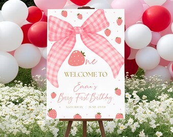 May include: A white welcome sign with a pink gingham bow and strawberries. The sign says "One Welcome to Emma's Berry First Birthday Saturday June 05th".