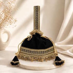 May include: Black velvet drawstring bag with gold embroidered trim and a matching handle. The bag features gold tassels and a decorative gold trim around the opening. The bag is sitting on a white marble surface.