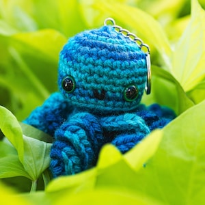 May include: A blue crocheted octopus keychain with large black eyes. The octopus is sitting in a bed of green leaves.