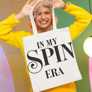 May include: A white tote bag with black text that reads "In My Spin Era". A person is holding the bag in front of their face.