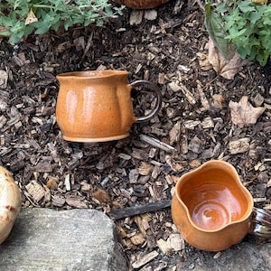 May include: Two orange ceramic mugs with a speckled finish and a dark brown handle. The mugs have a rounded, pumpkin-like shape with vertical indentations along the sides. One mug is shown from the side, while the other is viewed from above.