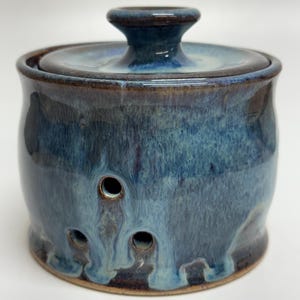 May include: A handmade ceramic garlic keeper with a lid. The jar is glazed in shades of blue and brown, with three circular holes near the base. The lid has a small handle. The jar is approximately 13 cm tall.