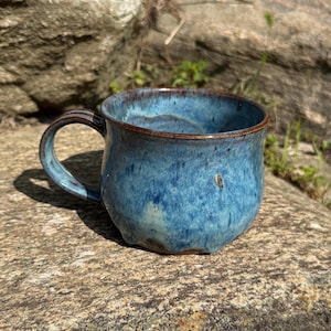 May include: A ceramic mug with a rounded body and a curved handle, featuring a mottled glaze in shades of deep blue, light blue, and hints of brown along the rim and handle. The surface has a glossy finish with subtle variations in color.