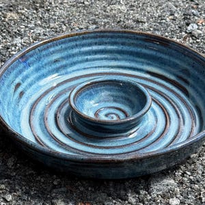 May include: A handmade ceramic serving bowl with a central dip bowl. The bowl features a spiral design in shades of blue and brown. The exterior has a dark brown rim. This pottery piece is ideal for serving snacks or dips.