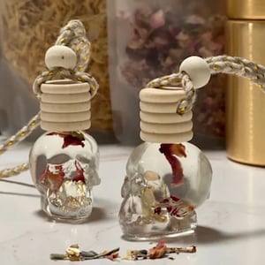 May include: Two clear glass skull-shaped air fresheners with wooden tops and decorative rope. The skulls are filled with dried flowers. The air fresheners are on a white surface.