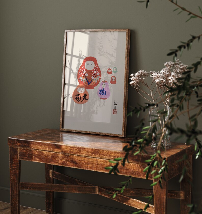 May include: A framed print featuring a collection of Daruma dolls in various sizes and colors. The largest doll is red with white floral accents, while others are purple, green, and orange. Japanese characters are visible on some of the dolls.