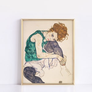 May include: Framed artwork featuring a seated figure with red hair, wearing a green top and black stockings. The artwork has a gold frame and is set against a white background. The style is reminiscent of early 20th-century expressionism.