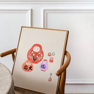 May include: A framed print featuring a collection of Japanese Daruma dolls in various sizes and colors. The dolls are primarily red, orange, and pink, with floral and character details. The print has Japanese text and is set against a cream background.