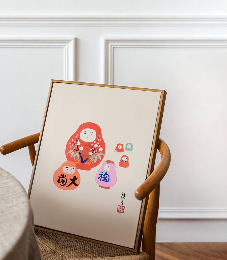 May include: A framed print featuring a collection of Japanese Daruma dolls in various sizes and colors. The dolls are primarily red, orange, and pink, with floral and character details. The print has Japanese text and is set against a cream background.