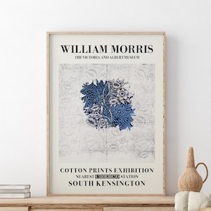 May include: Framed art print featuring a floral design in shades of blue and white, with the text "WILLIAM MORRIS" and "COTTON PRINTS EXHIBITION." The print is displayed in a light-colored wooden frame, resting on a wooden surface.