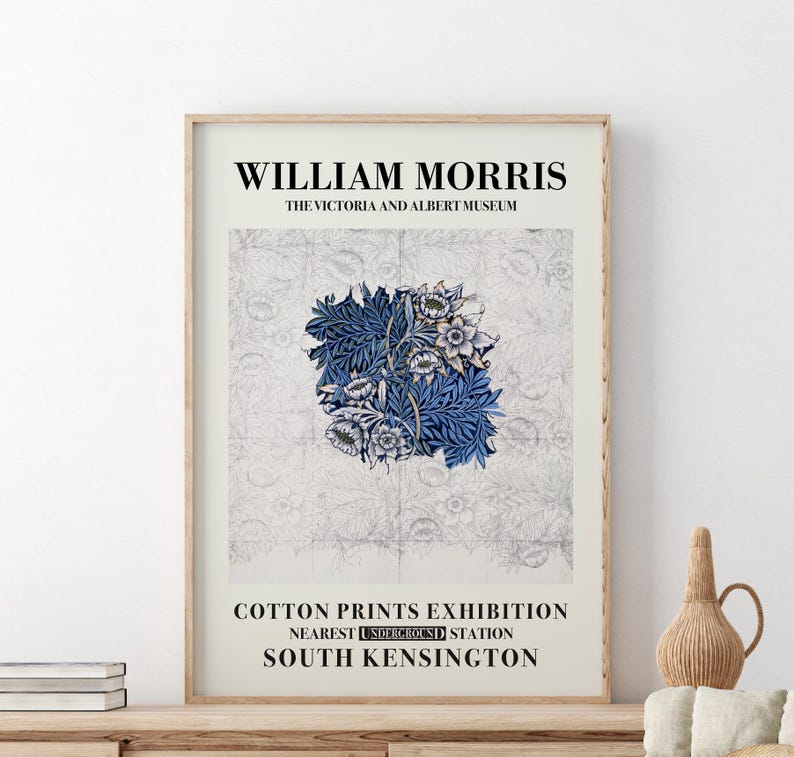 May include: Framed art print featuring a floral design in shades of blue and white, with the text "WILLIAM MORRIS" and "COTTON PRINTS EXHIBITION." The print is displayed in a light-colored wooden frame, resting on a wooden surface.