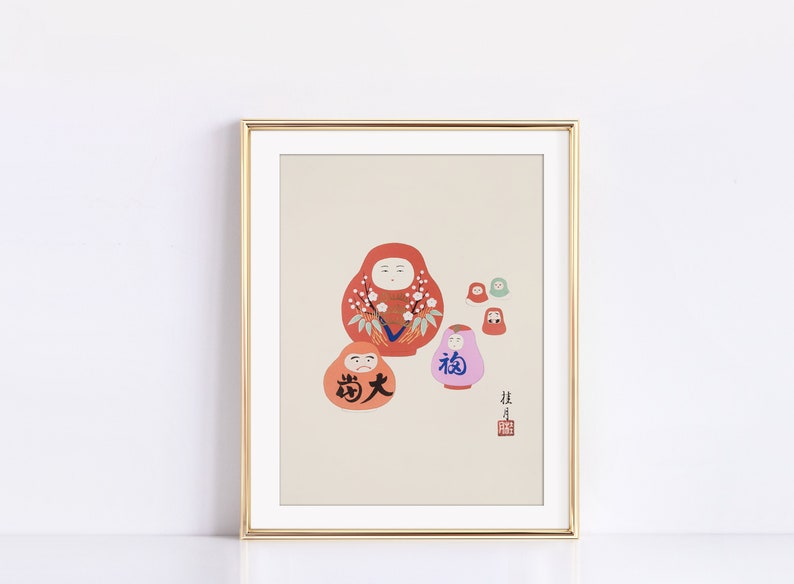 May include: Framed art print featuring a collection of Daruma dolls in shades of red, orange, and pink, with Japanese characters. The dolls have white facial features and floral accents. The print is in a gold frame.