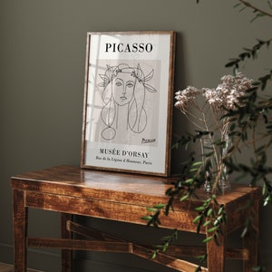 Puede incluir: Una impresi&oacute;n art&iacute;stica enmarcada con un dibujo lineal del rostro de una mujer, con el nombre "PICASSO" arriba y "MUS&Eacute;E D'ORSAY" abajo. La impresi&oacute;n est&aacute; en un marco de madera y descansa sobre una mesa de madera. La obra de arte es en tonos negros y beige.