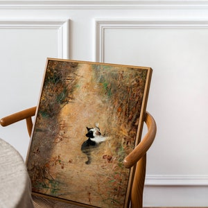 K&ouml;nnte beinhalten: Ein gerahmtes Kunstwerk mit einer Katze in einer nat&uuml;rlichen Umgebung. Das Gem&auml;lde zeigt eine schwarz-wei&szlig;e Katze inmitten einer Landschaft in warmen Erdt&ouml;nen, mit einem Holzrahmen. Das Kunstwerk wird auf einem Holzstuhl pr&auml;sentiert.