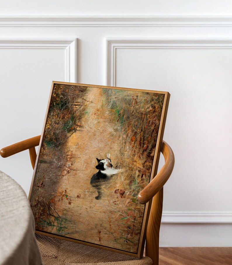 K&ouml;nnte beinhalten: Ein gerahmtes Kunstwerk mit einer Katze in einer nat&uuml;rlichen Umgebung. Das Gem&auml;lde zeigt eine schwarz-wei&szlig;e Katze inmitten einer Landschaft in warmen Erdt&ouml;nen, mit einem Holzrahmen. Das Kunstwerk wird auf einem Holzstuhl pr&auml;sentiert.