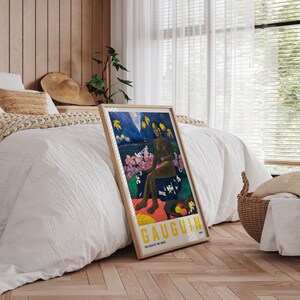 May include: A framed art print of Gauguin's "The Seed of the Areoi" featuring a seated figure in a tropical landscape. The artwork has a wooden frame and is propped against a bed with white bedding. The print includes yellow text.