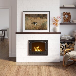 K&ouml;nnte beinhalten: Ein gem&uuml;tliches Wohnzimmer mit einem wei&szlig;en Ziegelkamin und einem gerahmten Bild dar&uuml;ber. Der Kamin hat ein brennendes Feuer. Ein h&ouml;lzerner Schaukelstuhl mit cremefarbenem Kissen steht daneben. Ein Teppich und Regale mit Dekoration vervollst&auml;ndigen das warme Interieur.