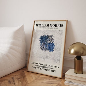 May include: A framed art print featuring a blue floral design, with the text "WILLIAM MORRIS" and "COTTON PRINTS EXHIBITION" in black. The print is displayed on a wooden floor, next to a gold lamp and a stack of books.