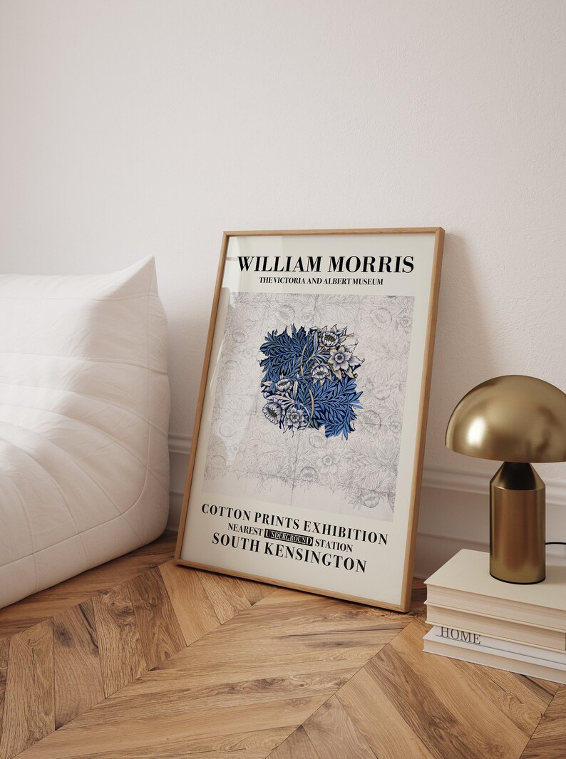 May include: A framed art print featuring a blue floral design, with the text "WILLIAM MORRIS" and "COTTON PRINTS EXHIBITION" in black. The print is displayed on a wooden floor, next to a gold lamp and a stack of books.