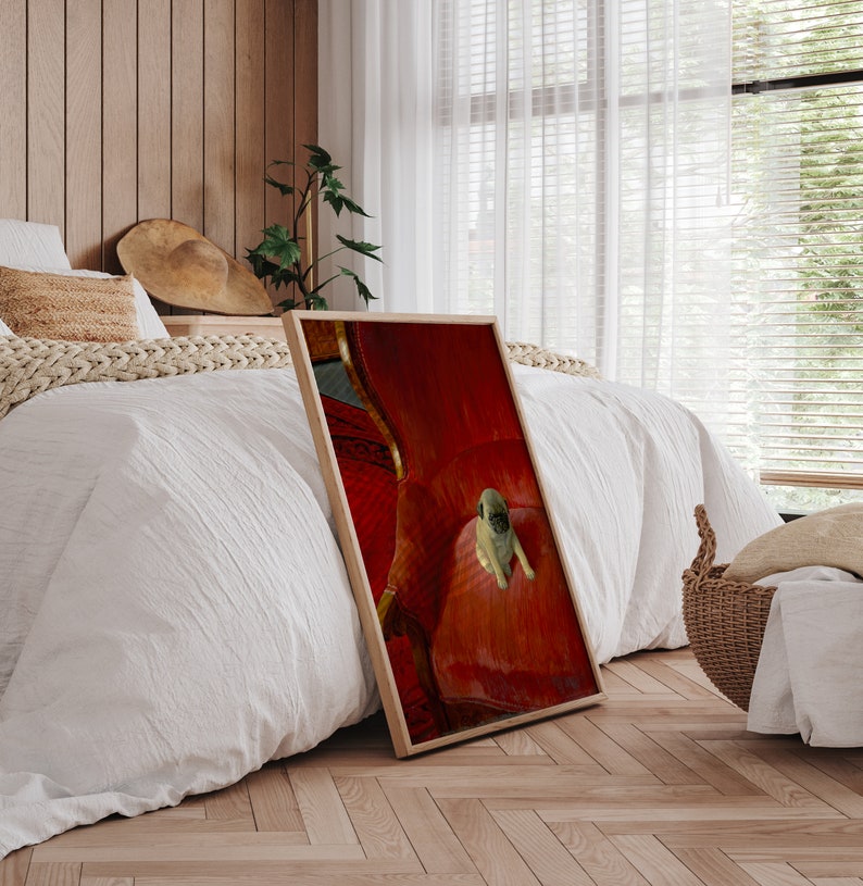 Puede incluir: Una impresi&oacute;n enmarcada que presenta un cachorro pug descansando sobre una silla roja tapizada. La impresi&oacute;n tiene un marco de madera y est&aacute; apoyada contra una cama con ropa de cama blanca. La habitaci&oacute;n tiene pisos de madera y una ventana con persianas.