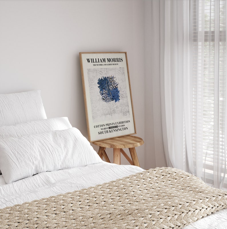 May include: A framed poster featuring the text "WILLIAM MORRIS THE VICTORIA AND ALBERT MUSEUM" and "COTTON PRINTS EXHIBITION SOUTH KENSINGTON." The poster has a floral design in blue and white. The poster is leaning against a wooden stool.