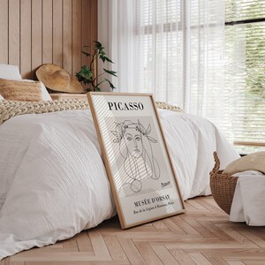 Puede incluir: Un cuadro enmarcado con un dibujo lineal de Picasso de un rostro femenino. La impresi&oacute;n tiene un fondo beige y est&aacute; etiquetada como "PICASSO" y "MUS&Eacute;E D'ORSAY". El marco es de color marr&oacute;n claro. La obra de arte se exhibe en un dormitorio.