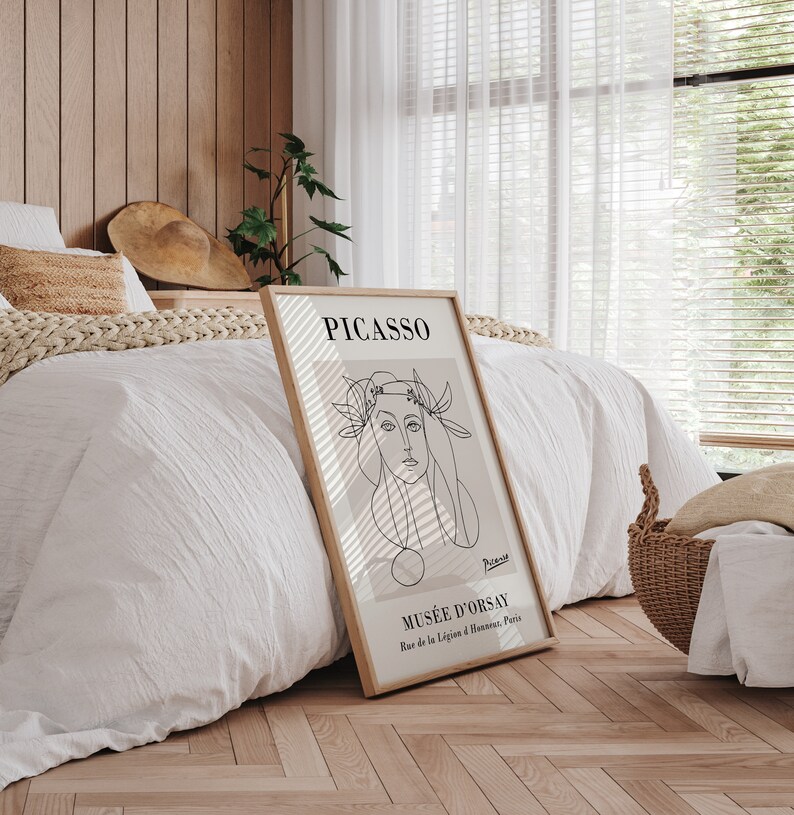 Puede incluir: Un cuadro enmarcado con un dibujo lineal de Picasso de un rostro femenino. La impresi&oacute;n tiene un fondo beige y est&aacute; etiquetada como "PICASSO" y "MUS&Eacute;E D'ORSAY". El marco es de color marr&oacute;n claro. La obra de arte se exhibe en un dormitorio.