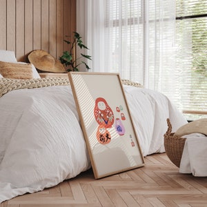 May include: A framed print featuring a Daruma doll illustration in shades of red, orange, and purple. The artwork has Japanese text and is set against a beige background with a light wooden frame. The print is leaning against a bed.