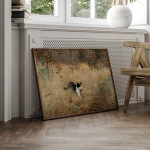 K&ouml;nnte beinhalten: Ein gerahmtes Gem&auml;lde zeigt eine schwarz-wei&szlig;e Katze auf einem Feld, die einen Vogel tr&auml;gt. Das Kunstwerk hat einen braunen Holzrahmen und lehnt an einer Wand. Die Szene spielt in einer nat&uuml;rlichen Umgebung mit hohem Gras und B&auml;umen.