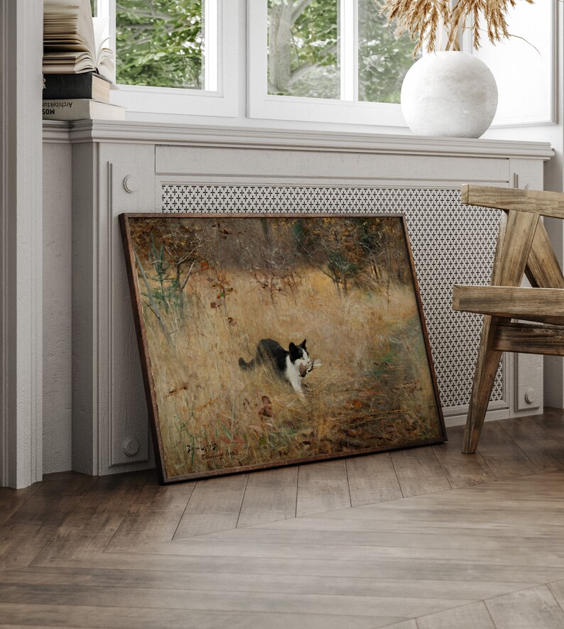 K&ouml;nnte beinhalten: Ein gerahmtes Gem&auml;lde zeigt eine schwarz-wei&szlig;e Katze auf einem Feld, die einen Vogel tr&auml;gt. Das Kunstwerk hat einen braunen Holzrahmen und lehnt an einer Wand. Die Szene spielt in einer nat&uuml;rlichen Umgebung mit hohem Gras und B&auml;umen.