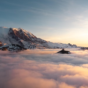 May include: A majestic mountain range with snow-capped peaks rises above a sea of clouds. The sky transitions from a soft blue to a warm golden hue, creating a serene and breathtaking landscape.